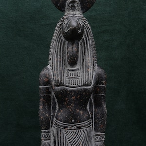 Puede incluir: Una estatua de granito negro de un dios egipcio con cuerpo humano y cabeza de chacal. El dios lleva un tocado egipcio tradicional y una falda. La estatua est&aacute; de pie sobre un fondo verde.