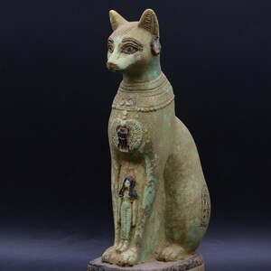 May include: Green painted ceramic statue of a seated cat with hieroglyphics on the base. The cat is wearing a necklace and has a small human figure carved on its chest.