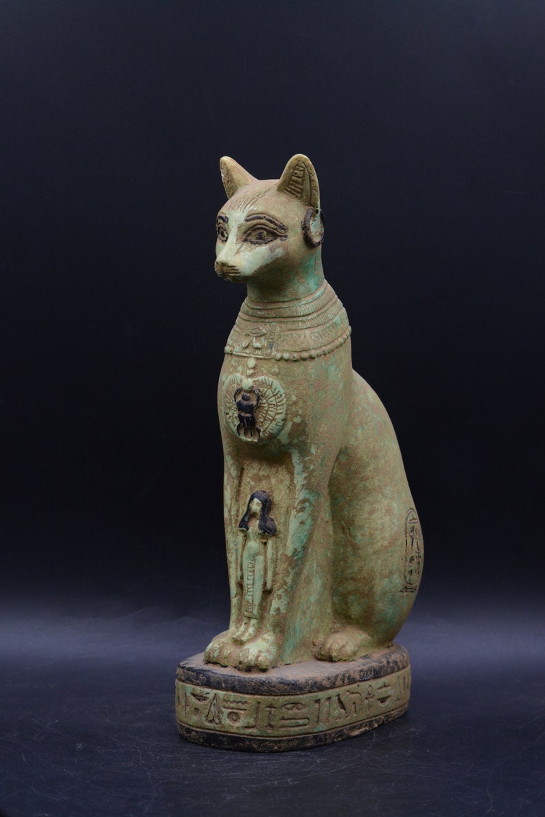 May include: Green painted ceramic statue of a seated cat with hieroglyphics on the base. The cat is wearing a necklace and has a small human figure carved on its chest.