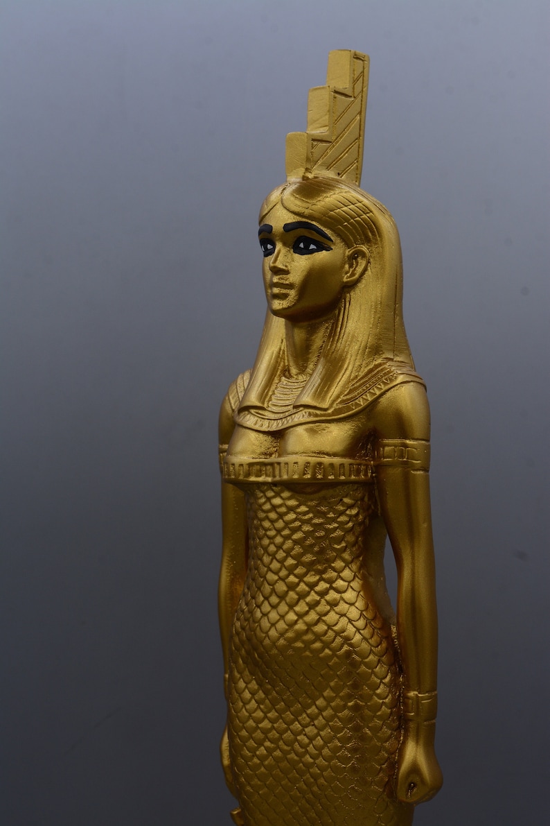 May include: A gold-coloured statue of a woman with long hair and a headdress. The statue features a detailed design, including a scaled dress and armbands. The figure is set against a grey background, highlighting the statue's intricate details and golden colour.