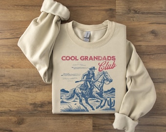 Może przedstawiać: Beżowa bluza z nadrukiem w stylu vintage przedstawiającym kowboja na koniu. Na przodzie bluzy znajduje się napis "COOL GRANDADS CLUB" w kolorze czerwonym i niebieskim.