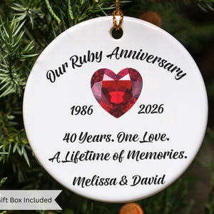 May include: A round white ceramic ornament featuring a red heart-shaped gemstone graphic. Text reads: Our Ruby Anniversary, 1986, 2026, 40 Years. One Love. A Lifetime of Memories. Melissa & David. The ornament has a gold hanging string at the top.