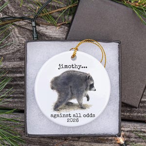 Pu&ograve; includere: Un ornamento rotondo in ceramica bianca con un'illustrazione dettagliata di un procione. Il testo "jimothy..." appare sopra il procione, mentre il testo "against all odds 2026" &egrave; stampato sotto. L'ornamento include un cordino dorato.