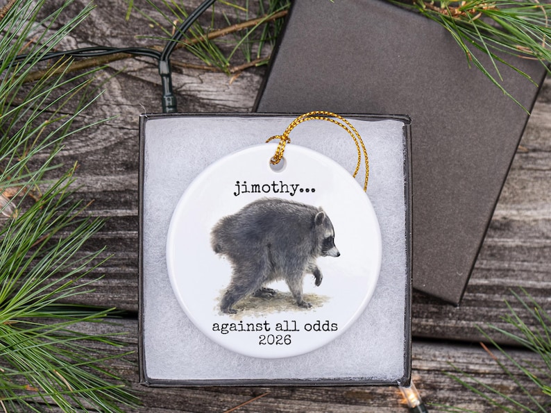 Pu&ograve; includere: Un ornamento rotondo in ceramica bianca con un'illustrazione dettagliata di un procione. Il testo "jimothy..." appare sopra il procione, mentre il testo "against all odds 2026" &egrave; stampato sotto. L'ornamento include un cordino dorato.