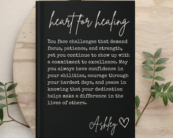 May include: A black hardcover book with white text that reads "heart for healing" followed by a paragraph about facing challenges and showing up with excellence. The text concludes with the name "Ashley" and a small heart symbol.