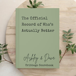 Può includere: Un libro segnapunti per cribbage con copertina rigida color verde salvia e testo nero che recita The Official Record of Who's Actually Better, seguito dai nomi Ashley & Dave in carattere corsivo e la parola Cribbage Scorebook in basso.