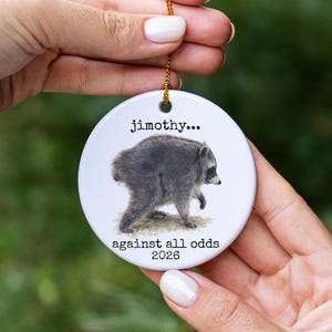 Pu&ograve; includere: Un ornamento rotondo in ceramica bianca con un'illustrazione dettagliata di un procione. Il testo "jimothy..." &egrave; stampato sopra il procione e il testo "against all odds 2026" &egrave; stampato sotto. Un cordino dorato &egrave; fissato al foro superiore.