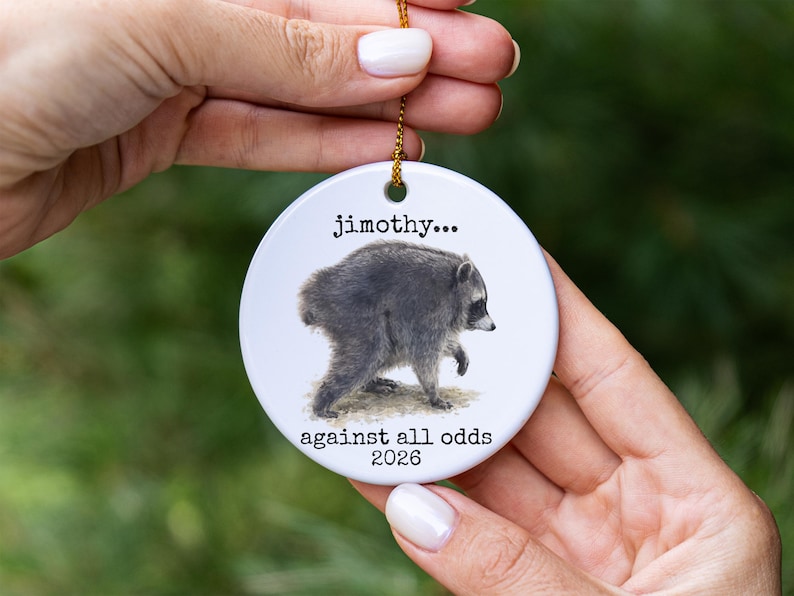 Pu&ograve; includere: Un ornamento rotondo in ceramica bianca con un'illustrazione dettagliata di un procione. Il testo "jimothy..." &egrave; stampato sopra il procione e il testo "against all odds 2026" &egrave; stampato sotto. Un cordino dorato &egrave; fissato al foro superiore.