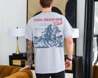 May include: Grey t-shirt featuring a graphic of a cowboy riding a horse. The text "Cool Grandpas Club" is printed on the shirt.