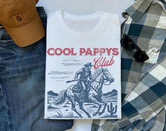 May include: White t-shirt featuring a cowboy on horseback graphic, with the text "COOL PAPPY'S Club" in red. The shirt is displayed with a brown cap, blue jeans, a plaid shirt, and sunglasses.