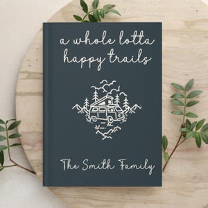 May include: A dark blue hardcover journal featuring the white text "a whole lotta happy trails" at the top and "The Smith Family" at the bottom. The center displays a white line art illustration of a camper van surrounded by trees and mountains.