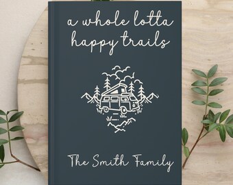 May include: A dark blue hardcover journal featuring the white text "a whole lotta happy trails" at the top and "The Smith Family" at the bottom. The center displays a white line art illustration of a camper van surrounded by trees and mountains.