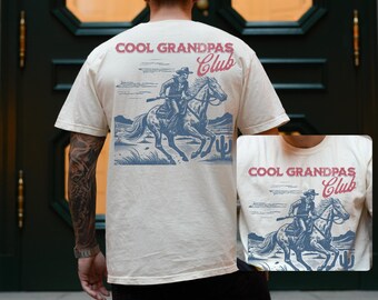 May include: A cream-coloured t-shirt featuring a blue and red graphic of a cowboy riding a horse, with the text "COOL GRANDPAS Club". The design includes a desert landscape. This is a casual wear item.