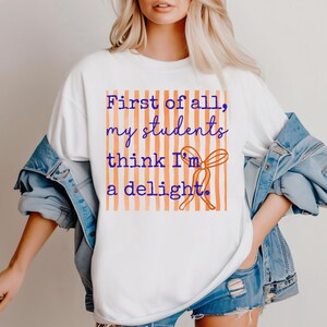 Może przedstawiać: Biała koszulka z napisem "First of all, my students think I'm a delight" w kolorze niebieskim i pomarańczowym. Tekst jest wyśrodkowany na prostokątnym, pomarańczowo-białym tle w paski. Dołączona grafika kokardy.