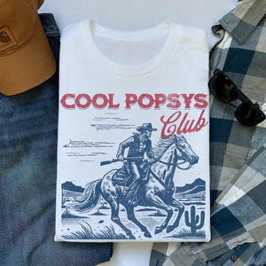 May include: White t-shirt with a blue and red graphic of a cowboy riding a horse. The graphic says "Cool Popsys Club".