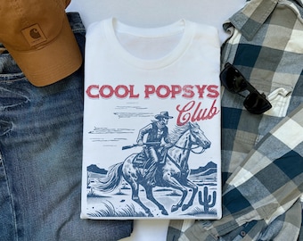 May include: White t-shirt with a blue and red graphic of a cowboy riding a horse. The graphic says "Cool Popsys Club".