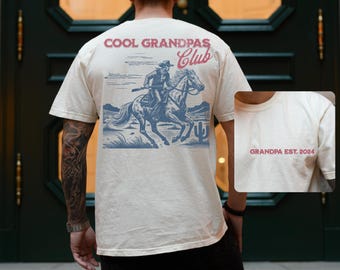 May include: A cream-coloured t-shirt featuring a cowboy on horseback graphic, with the text "COOL GRANDPAS Club" and "GRANDPA EST. 2024". The design is in blue and red. This is a casual wear t-shirt.