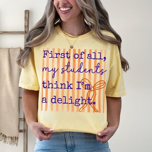 Może przedstawiać: Jasnoż&oacute;łta koszulka z napisem "First of all, my students think I'm a delight" w niebieskim skrypcie. Projekt zawiera pomarańczowe pionowe paski i pomarańczową kokardę. Koszulka jest noszona przez osobę.