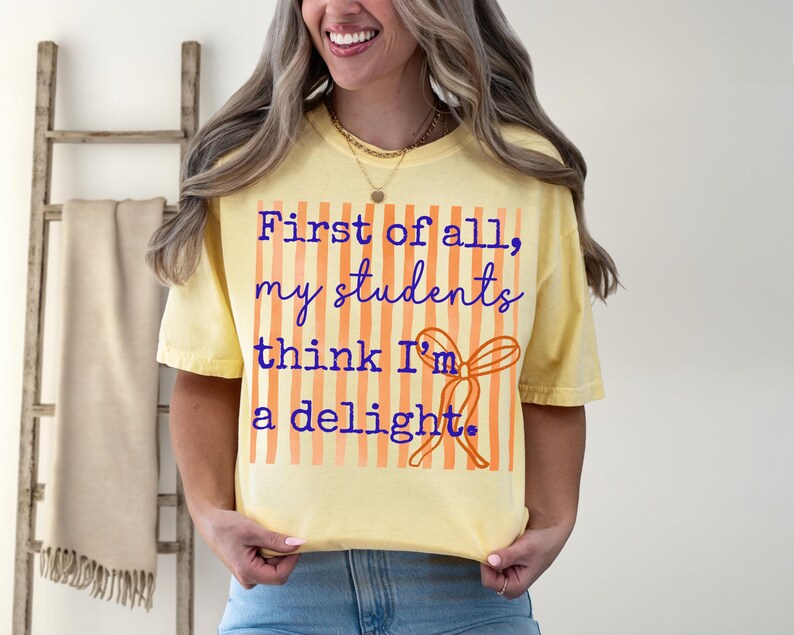 Może przedstawiać: Jasnoż&oacute;łta koszulka z napisem "First of all, my students think I'm a delight" w niebieskim skrypcie. Projekt zawiera pomarańczowe pionowe paski i pomarańczową kokardę. Koszulka jest noszona przez osobę.