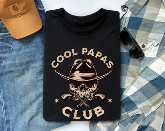 May include: Black t-shirt featuring a graphic of a skull wearing a cowboy hat and holding two pistols. The text "COOL PAPAS CLUB" is written around the skull.