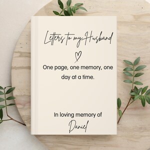 May include: A cream-coloured hardback book with black text that reads Letters to my Husband, a heart symbol, One page, one memory, one day at a time., and In loving memory of Daniel. The book is centred on a round, light-coloured stone surface.