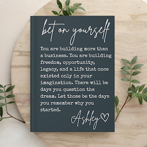 May include: A dark gray hardcover journal featuring the white text "bet on yourself" followed by an inspirational paragraph about building a business and legacy, ending with the name "Ashley" and a heart symbol printed on the front cover.