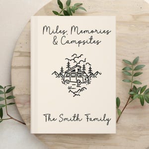 May include: A cream-colored hardcover book featuring black line art of a camper van surrounded by trees and mountains. The text reads Miles, Memories & Campsites at the top and The Smith Family at the bottom in a simple black script font.
