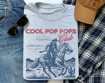 May include: A gray crew neck t-shirt with the text "COOL POP POPS Club" in red and a blue graphic of a cowboy riding a horse. The shirt is surrounded by blue jeans, sunglasses, a brown cap, and a plaid shirt.