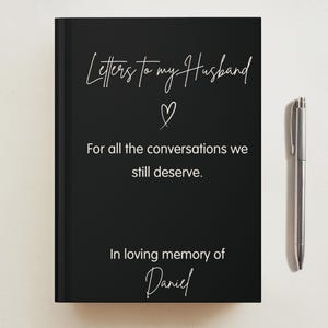 May include: A black hardback journal with white text that reads Letters to my Husband, a heart icon, For all the conversations we still deserve., and In loving memory of Daniel. A silver metal pen is positioned vertically to the right of the journal.
