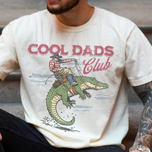 Op de afbeelding: Een crèmekleurig T-shirt met korte mouwen en een grafische print van een persoon met een cowboyhoed en geruit overhemd die op een groene alligator rijdt. De tekst COOL DADS Club staat in het rood boven de illustratie met een lasso en lijnen.