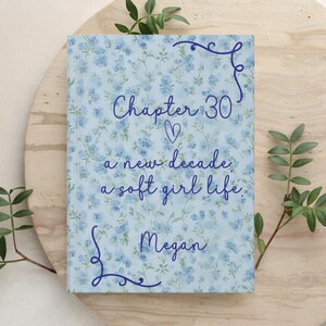 May include: A floral-patterned book cover with the text "Chapter 30, a new decade, a soft girl life, Megan" in blue script. The cover features a light blue background with small blue flowers. Decorative flourishes frame the text.