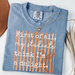 Może przedstawiać: Niebieska koszulka Comfort Colors z napisem "First of all, my students think I'm a delight" w kolorze białym i pomarańczowym. Złoty naszyjnik z wisiorkiem jest zarzucony na koszulkę.
