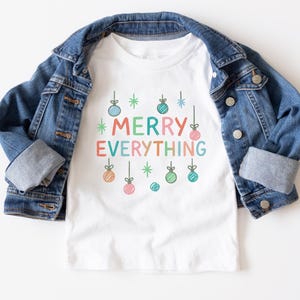Pode incluir: Uma camiseta branca com a frase "MERRY EVERYTHING" em letras coloridas e ilustrações de enfeites de Natal. Uma jaqueta jeans azul está sobre a camiseta.