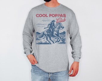 May include: A grey long-sleeved shirt featuring a cowboy on horseback graphic, with the text "COOL POPPAS Club" in red and blue. The cowboy is holding a rifle. The shirt is made of a soft material.