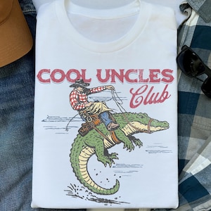 May include: A white t-shirt featuring a graphic print of a person in a red plaid shirt and cowboy hat riding a green alligator. The text above the graphic reads COOL UNCLES Club in red and maroon lettering. The design has a distressed texture.