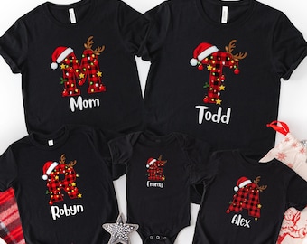 May include: A collection of black t-shirts and a black baby bodysuit featuring a red and black buffalo plaid letter graphic adorned with a Santa hat, reindeer antlers, and Christmas lights, with the names Mom, Todd, Robyn, Emma, and Alex.