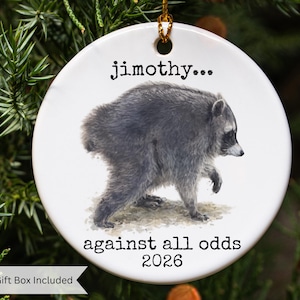 Pu&ograve; includere: Un ornamento rotondo in ceramica bianca con un'illustrazione dettagliata di un procione che cammina. Il testo "jimothy..." &egrave; stampato sopra il procione, mentre il testo "against all odds 2026" &egrave; riportato sotto. Un filo dorato &egrave; fissato in alto.
