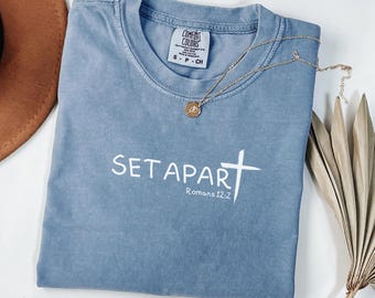 Può includere: T-shirt blu a maniche corte con le parole bianche "SET APART" e una croce. Il testo "Romani 12:2" è sotto le parole. Una collana d'oro con un ciondolo è sulla maglietta.
