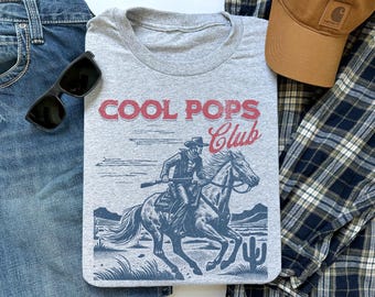 May include: A gray crew neck t-shirt with the words "COOL POPS Club" in red. The shirt features a graphic of a cowboy riding a horse. Sunglasses, a brown cap, and blue jeans are also in the image.