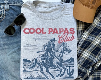 May include: A grey t-shirt featuring a vintage-style illustration of a cowboy riding a horse. The text "COOL PAPAS Club" is printed in red and blue on the shirt.