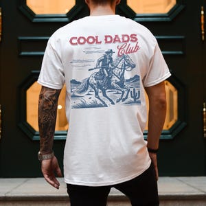 May include: White t-shirt with a graphic of a cowboy riding a horse. The graphic is in blue and red and says "Cool Dads Club".