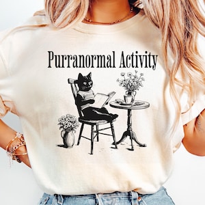Puede incluir: Camiseta color crema con el texto negro "Purranormal Activity". El gráfico muestra un gato negro leyendo un libro, sentado en una silla, con una pequeña mesa y flores. El diseño es una versión caprichosa de un tema clásico.