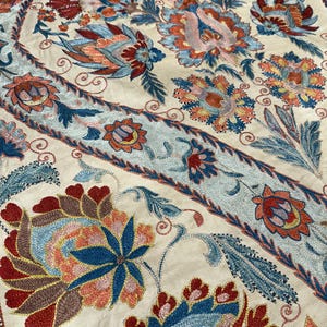 Op de afbeelding: Crèmekleurig textiel met ingewikkelde bloemenborduurwerk in rood, blauw, oranje en geel. Het ontwerp bevat grote, gestileerde bloemen en bladeren, met gedetailleerde steken. Het borduurwerk is omzoomd door een lichtblauwe band.