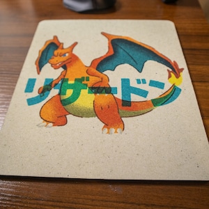 May include: A brown and orange cartoon character with blue wings and a fiery tail. The character is standing on a beige mousepad with the Japanese text "リザードン" printed in green, blue, and yellow.