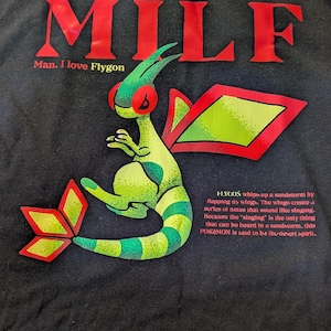Puede incluir: Camiseta negra con un gráfico rojo "MILF" y un dibujo animado verde y amarillo de Flygon Pokemon. El texto "Man. I love Flygon" está debajo del gráfico. El texto "FLYCON whips up a sandstorm by flapping its wings. The wings create a series of notes that sound like singing. Because the "singing" is the only thing that can be heard in a sandstorm, this POKEMON is said to be the desert spirit." está debajo del Pokemon.