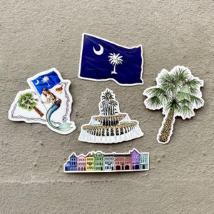May include: Five colorful stickers featuring various Charleston, South Carolina landmarks. The stickers include a mermaid, a fountain, a palm tree, a row of colorful houses, and the South Carolina state flag.