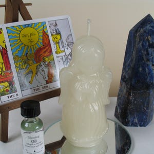 May include: An off-white angel-shaped candle, tarot cards, a blue crystal point, and a small bottle of oil are arranged on a mirrored surface. The tarot cards are displayed on a small wooden easel. The Sun card is visible.