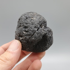 Peut inclure: Une roche de couleur gris foncé, rugueuse et texturée, avec une forme légèrement arrondie. La roche a un aspect naturel et organique.