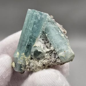May include: A raw aquamarine crystal cluster. The crystal is a pale blue-green color, with a translucent appearance. The crystal is partially embedded in a matrix of grey and white rock. The overall shape is a triangular prism.