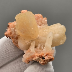 Peut inclure: Un amas de cristaux couleur pêche avec une teinte crème et blanc cassé. La formation minérale a un aspect rugueux et texturé, avec des cristaux allongés et d'autres plus arrondis. La base de l'amas est de couleur brun rougeâtre.